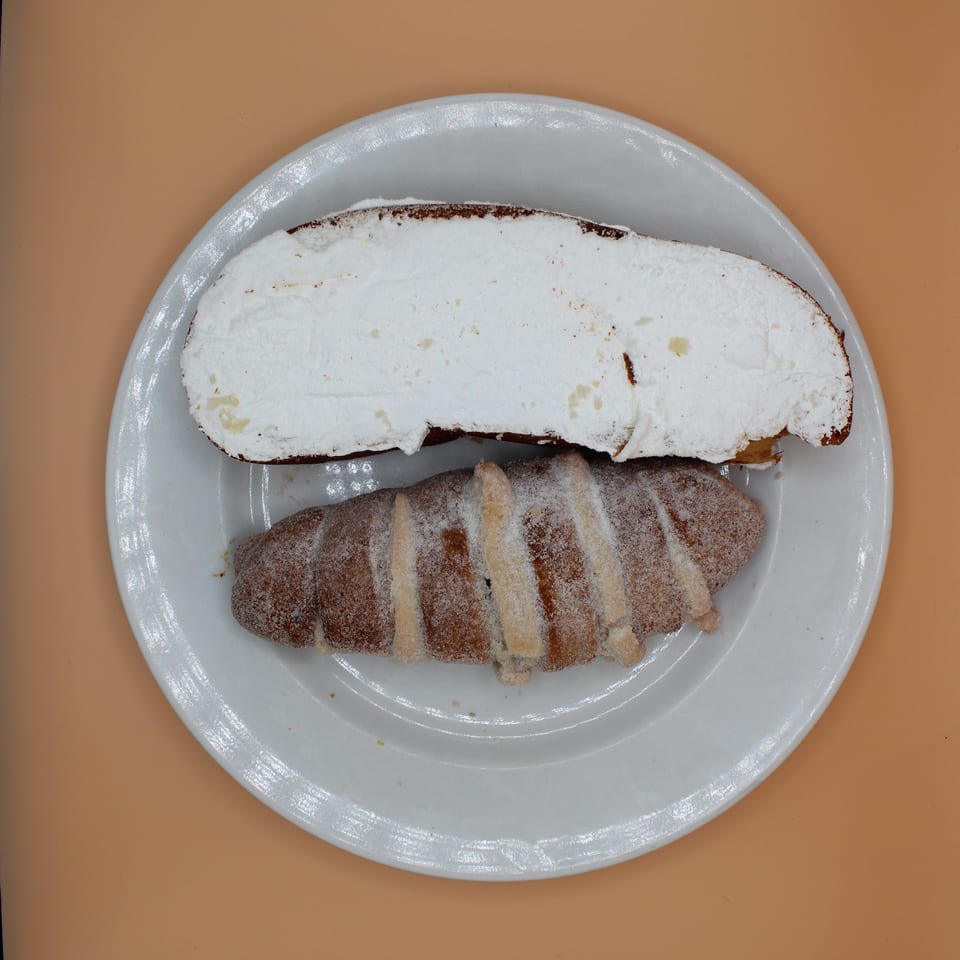 A white plate holds a croissant topped with whipped cream and a sugar-dusted pastry.